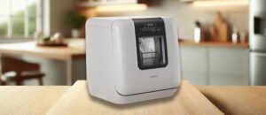 A featured image of the topic COMFEE CDC17P2AWW Dishwasher Review or COMFEE MINIPLUS CDC17P2AWW Dishwasher Review – A modern, compact countertop dishwasher in a sleek white finish, placed on a wooden kitchen counter. The dishwasher features a small front window showing neatly arranged dishes inside, along with a digital control panel on the top front. The cozy kitchen setting in the background enhances its aesthetic appeal and practical design for small spaces.