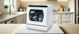 A featured image of the topic Hermitlux HMX-TDJ03 Dishwasher Review. Compact white Hermitlux HMX-TDJ03 countertop dishwasher displayed on a light wooden counter in a bright, modern kitchen. The dishwasher is shown with clean dishes inside, including glasses and plates, and features a digital display and touch controls on top.