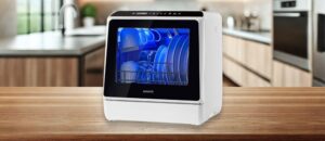 A featured image of the topic NOVETE TDQR01 Dishwasher Review. Compact white NOVETE TDQR01 countertop dishwasher placed on a wooden kitchen counter, displaying clean dishes inside with a glowing blue interior light. Modern kitchen setting in the background.