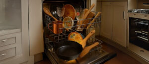 Open dishwasher loaded with items like cast iron, wooden utensils, and copper mugs, illustrating examples from the list of 21 things you should never put in the dishwasher.