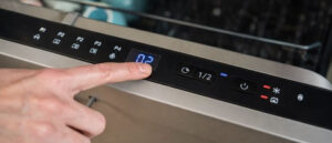Dishwasher Control Panel Is Not Working shown in a wide close-up of a modern stainless steel dishwasher, with a hand pressing the digital control panel buttons and illuminated display indicating control issues.