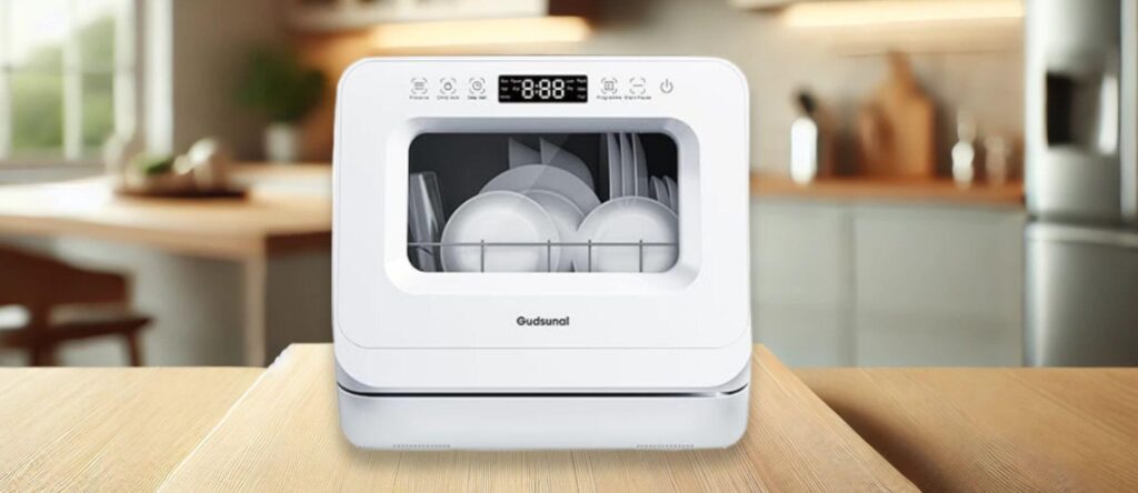 A lifestyle image showing the Gudsunal WQP4 6207 dishwasher placed on a wooden kitchen counter with plates and glasses inside, emphasizing its compact design and transparent window.