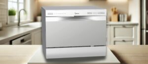 A high-resolution image of the Midea MDC22P2ASS dishwasher placed on a kitchen countertop. The background features a modern kitchen with white cabinets, a sink, and green plants. The dishwasher has a sleek stainless steel design with a control panel on the top front edge. It is a featured image of the topic "Midea MDC22P2ASS dishwasher review"