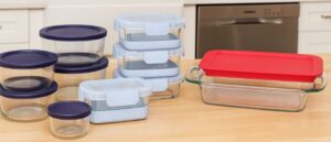 A variety of the best dishwasher-safe glass food containers displayed neatly on a bright kitchen counter, featuring round and rectangular shapes with navy, light blue, and red lids, with a built-in dishwasher in the background.