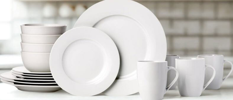 Amazon Basics 16-piece porcelain dinnerware set with plates, bowls, and mugs, arranged neatly on a bright kitchen surface, all safe for dishwashers.