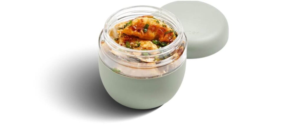 A single W&P Porter glass lunch bowl with a sage green silicone base, filled with dumplings and topped with herbs.