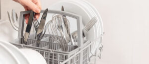 Close-up of a dishwasher cutlery basket filled with forks, knives, and a whisk, with a hand placing utensils beside clean white plates in the dishwasher rack