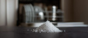 Close-up of dishwasher salt crystals on a dark counter with an open dishwasher in the background.