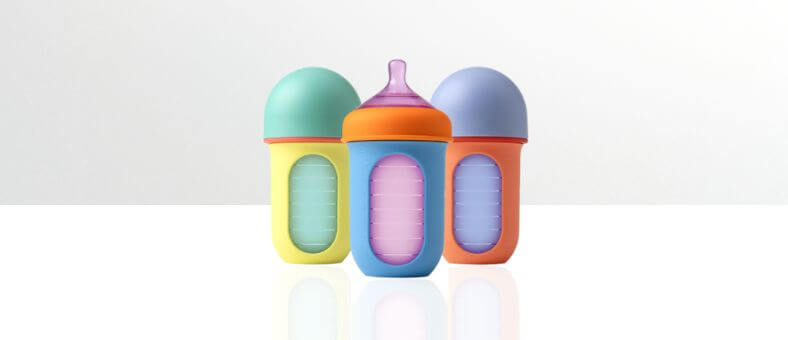 Boon Nursh Baby Bottles – Three colorful silicone baby bottles in blue, orange, and yellow, each with a transparent measurement window, displayed on a white reflective surface.