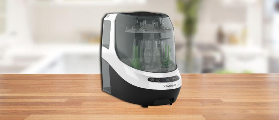 In this Baby Brezza Baby Bottle Washer Review image, the washer is displayed on a wooden countertop in a bright kitchen, showcasing its sleek black-and-white design with transparent lid and digital control panel.