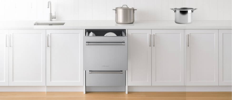 A bright, modern kitchen showcases the FIsher & Paykel DD24DV2T9N dishwasher built seamlessly into white cabinetry, with the top DishDrawer slightly open to reveal neatly arranged dishes. The stainless steel finish blends smoothly with the clean countertop, where two cooking pots rest near the sink. This scene highlights the stylish, functional design discussed in the Fisher & Paykel DD24DV2T9N dishwasher review.