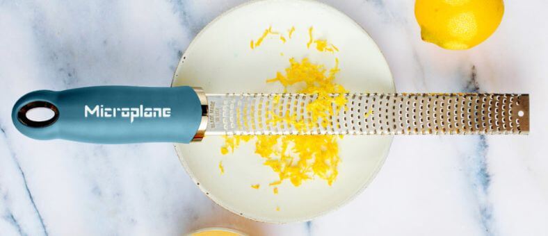 Microplane blue-handle zester grating fresh lemon zest over a plate.