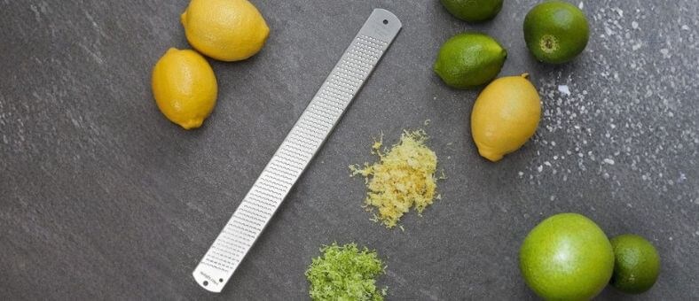 Stainless steel lemon zester surrounded by lemons, limes, and piles of zest.